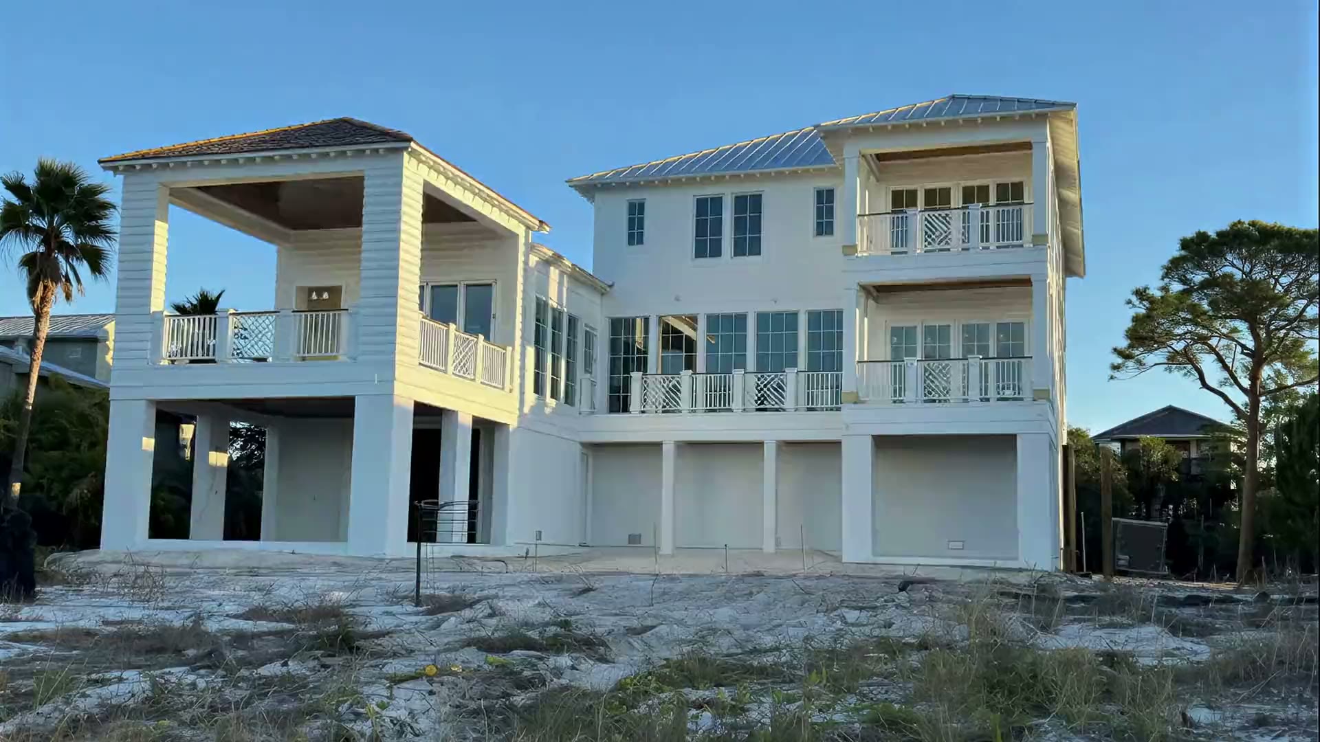 A white three-storey beach house on pilings with porches and white railings, seen across sand dunes.
