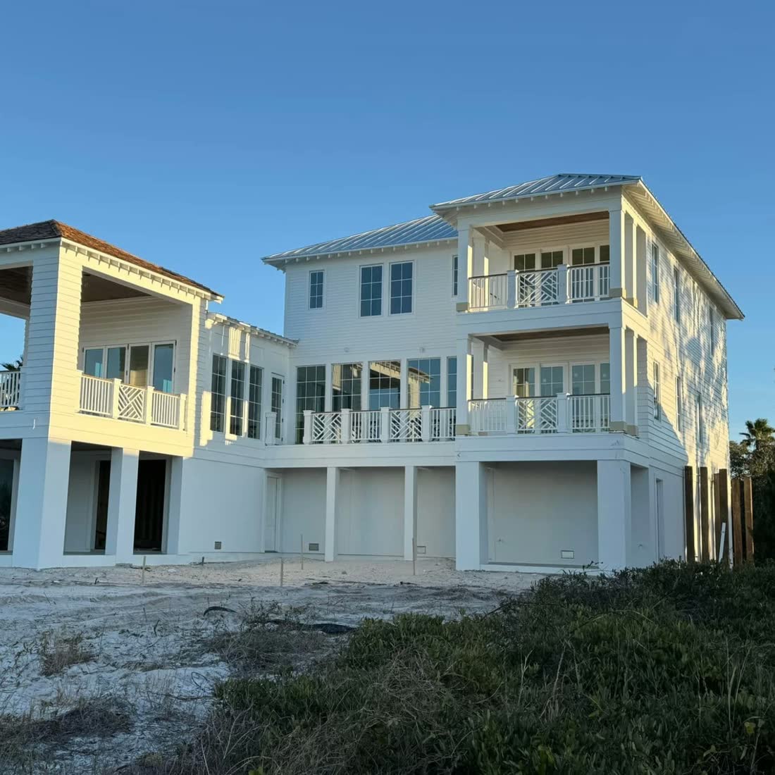 A large white raised beach house with stacked porches and white railings, seen from the beach at sunset.