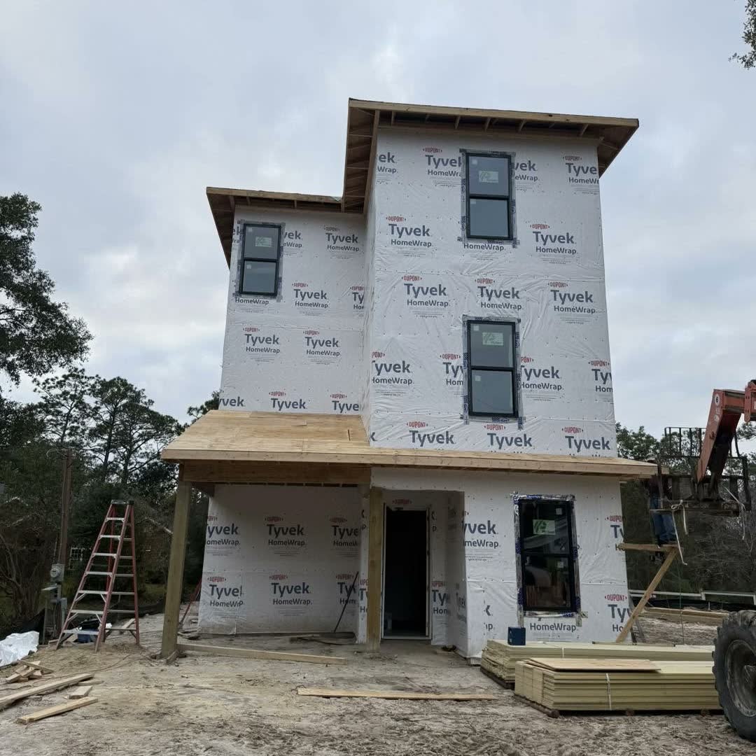 A new three-storey house in white house wrap with its black-framed windows in, on a sandy building lot.