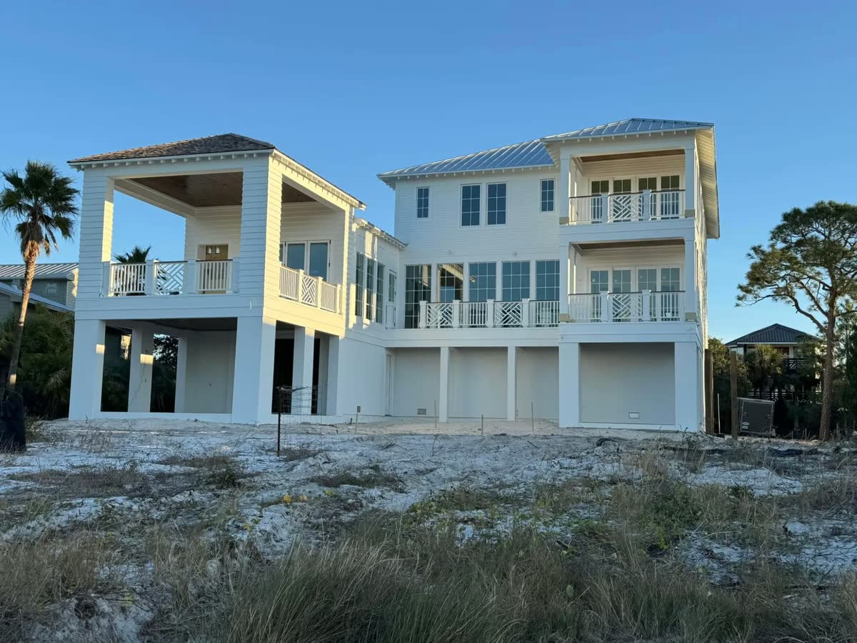 A white three-storey beach house with porches and white railings, seen across sand dunes.