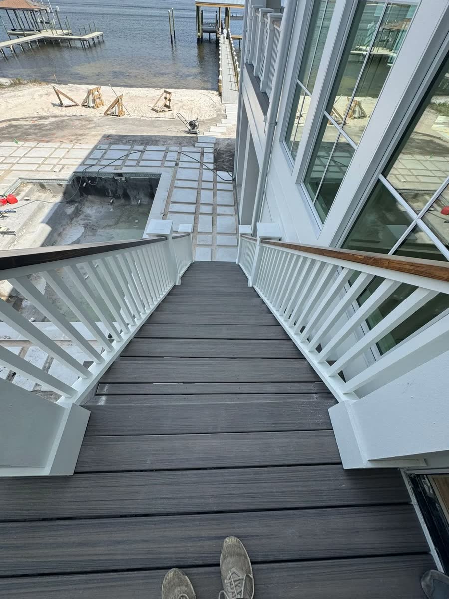 Looking down a stair with white railings toward a new pool deck and a pier on the bay.