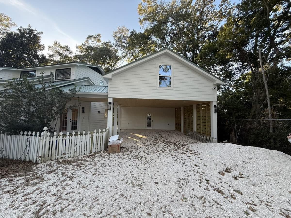 A white two-storey house with a carport underneath, a white picket fence and a sandy yard.