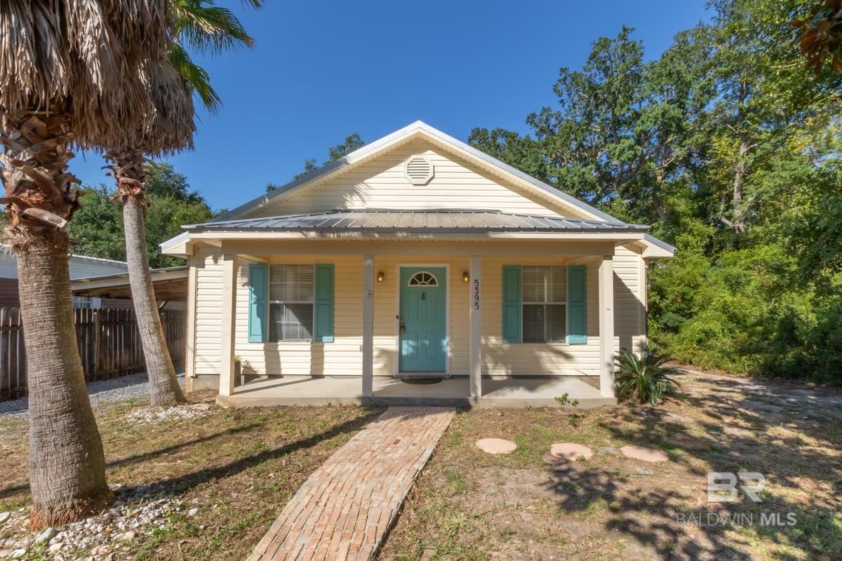 A pale yellow cottage with teal shutters and a teal front door, a brick path to the porch.