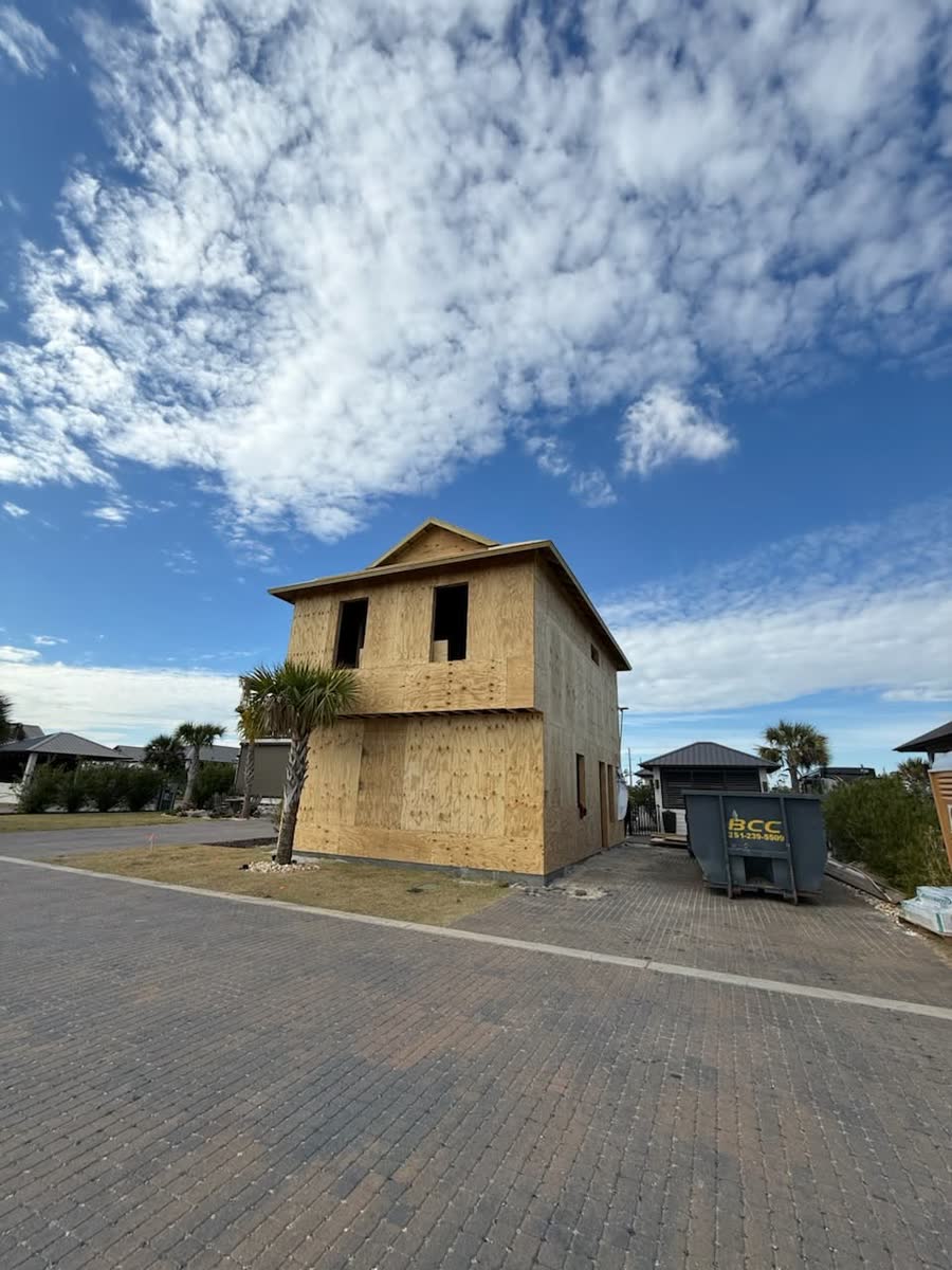 A small two-storey house sheathed in plywood beside the street, a dumpster at the curb.