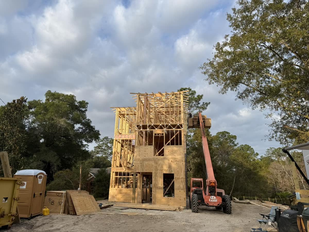 A three-storey house framed in raw lumber among trees, a telehandler beside it.