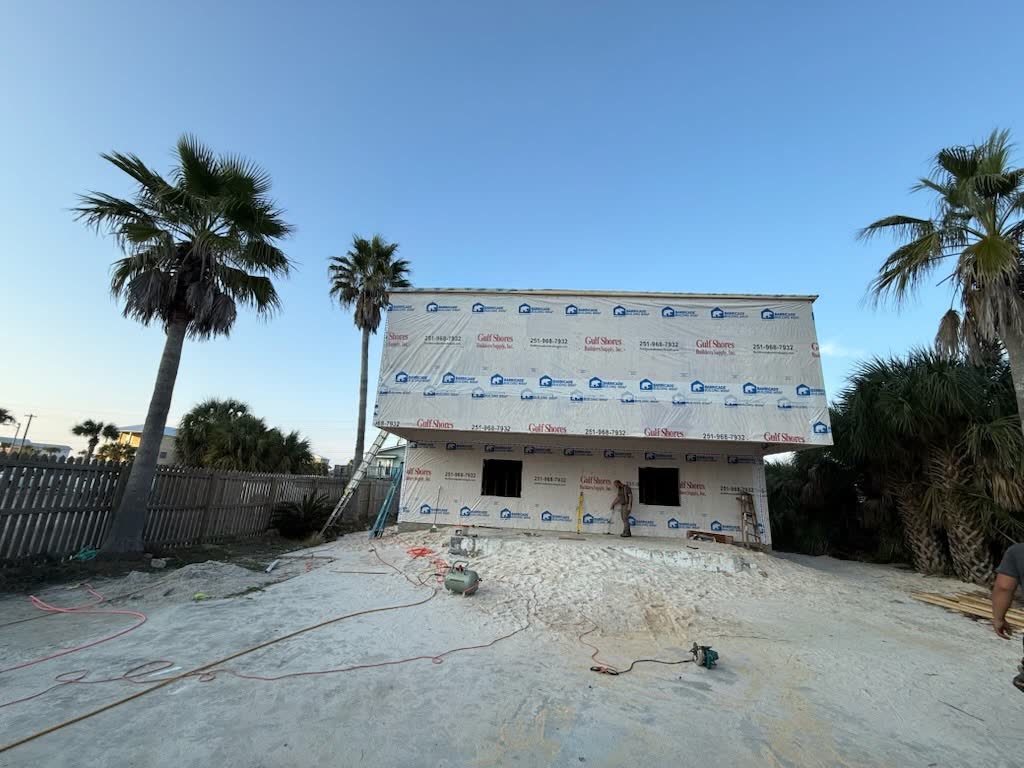 A raised house wrapped in house wrap on a sandy lot between palm trees.