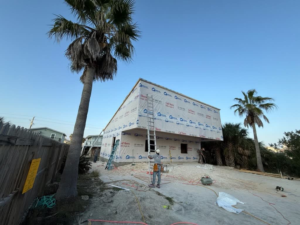 The side of a raised house in house wrap, a worker at the foot of it, palms behind.