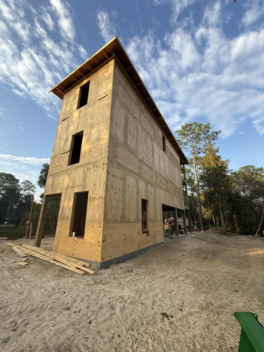 A tall narrow house sheathed in plywood on a sandy lot.
