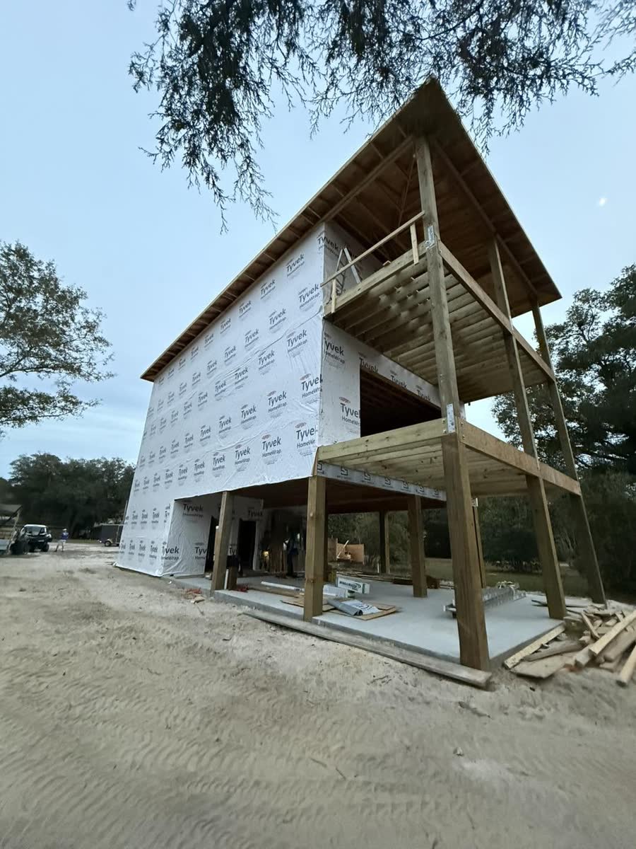 A raised house sheathed and covered in white house wrap, its porches framed in wood.