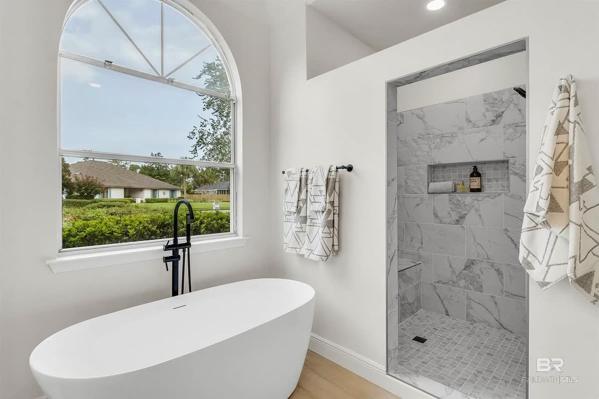 A remodeled bathroom with a freestanding tub, a veined-tile shower and an arched window.